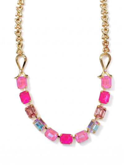Ribbon of Hope Tennis Necklace - NGM2BGPIN - <p>A structured sweep of pink emerald-cut crystals frames the collarbone, held by sculpted gold-tone ribbon motifs. The stones transition through shades of soft rose and electric pink, punctuated by flashes of iridescence that catch ambient light. At the back, an open-link chain balances the profile, allowing the brightly colored center to take focus. The adjustable length lets you place the stones exactly where you want them, whether layered over a collar or framing bare skin.</p><ul>
<li>Emerald-cut crystals in shifting pink and iridescent tones</li>
<li>Sculpted metal ribbon motifs anchor an open-link chain</li>
<li>Lobster claw clasp</li>
<li>4-inch adjustable extension chain</li>
<li>Protective e-coat finish for lasting wear</li>
<li>Includes organza pouch for gifting and travel</li>
</ul>